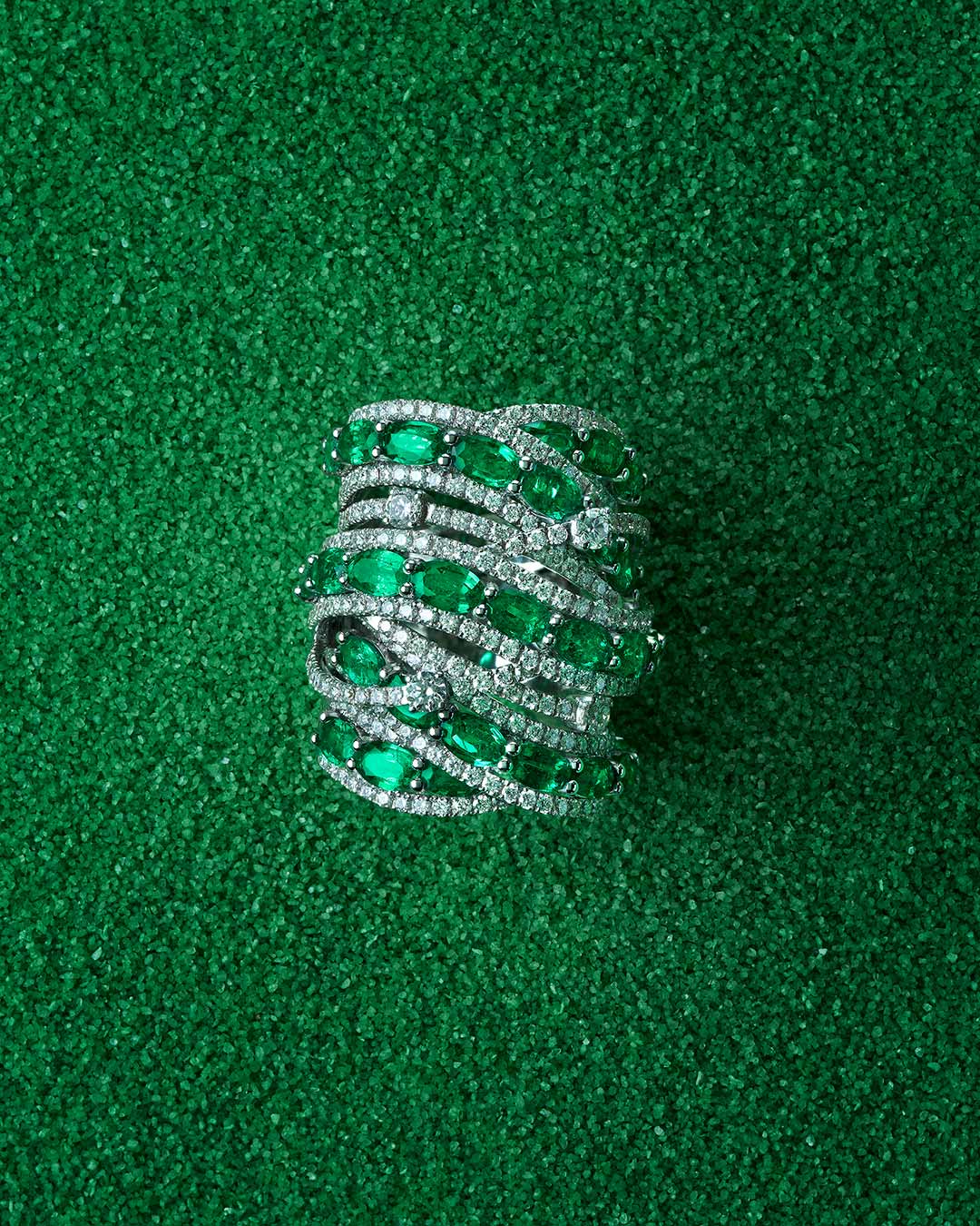 Anillo de alta joyería sobre fondo verde texturizado. La pieza presenta  esmeraldas entrelazadas con diamantes blancos engastados . El diseño combina brillo y color con una silueta sofisticada y orgánica.
