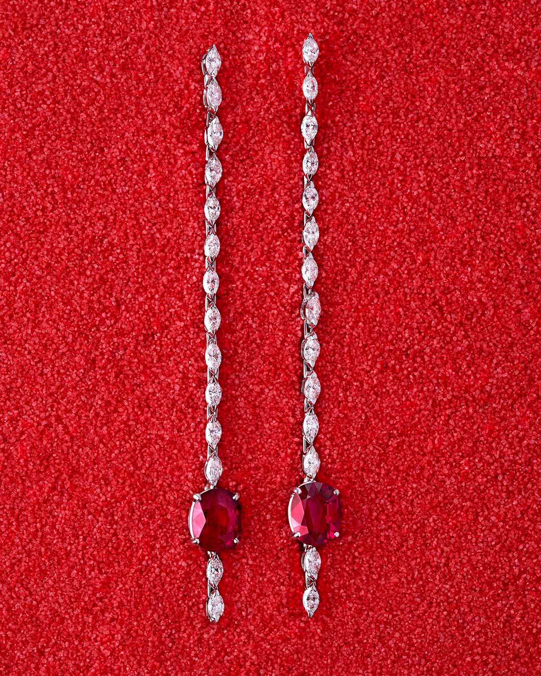 Pendientes largos sobre fondo rojo texturizado. Cada pieza presenta una línea de diamantes blancos que culmina en un rubí central de intenso rojo. El contraste entre el brillo de las piedras y la profundidad del fondo realza la sofisticación de la joya.
