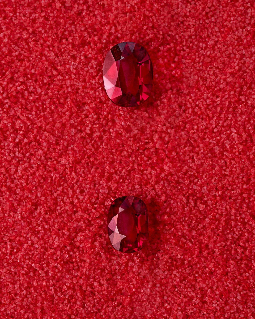 Dos rubíes rojos de talla oval sobre fondo rojo texturizado. Ambas piedras están facetadas y reflejan la luz con intensidad. La superior es ligeramente más alargada que la inferior. El contraste entre el acabado pulido de las gemas y la superficie granulada crea una composición visualmente armoniosa y sofisticada.