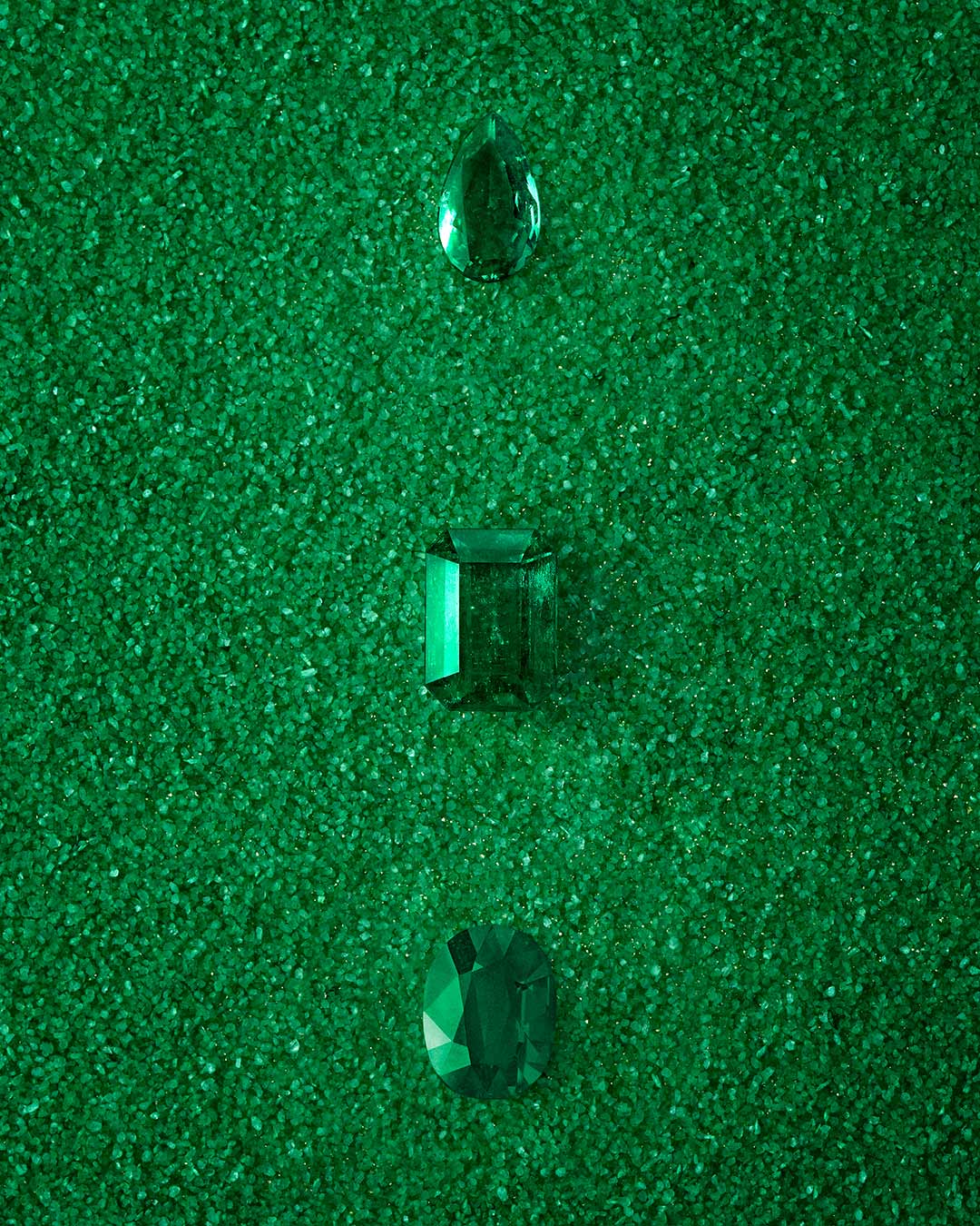 Tres esmeraldas verdes sobre fondo verde texturizado. Cada esmeralda presenta un corte distinto: lágrima, esmeralda y brillante redondo. La composición monocromática resalta la pureza del color y la precisión de cada forma, creando una imagen armoniosa y sofisticada.