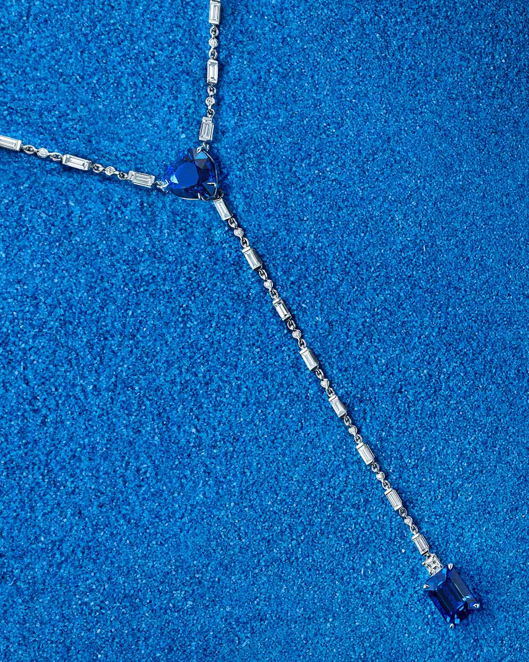 Collar sobre fondo azul texturizado. La pieza combina eslabones metálicos plateados con dos zafiros azules: un zafiro en forma de corazón en el centro del diseño y otra rectangular al final de la cadena. El contraste entre el brillo del metal y la profundidad del azul crea una composición refinada y visualmente impactante.