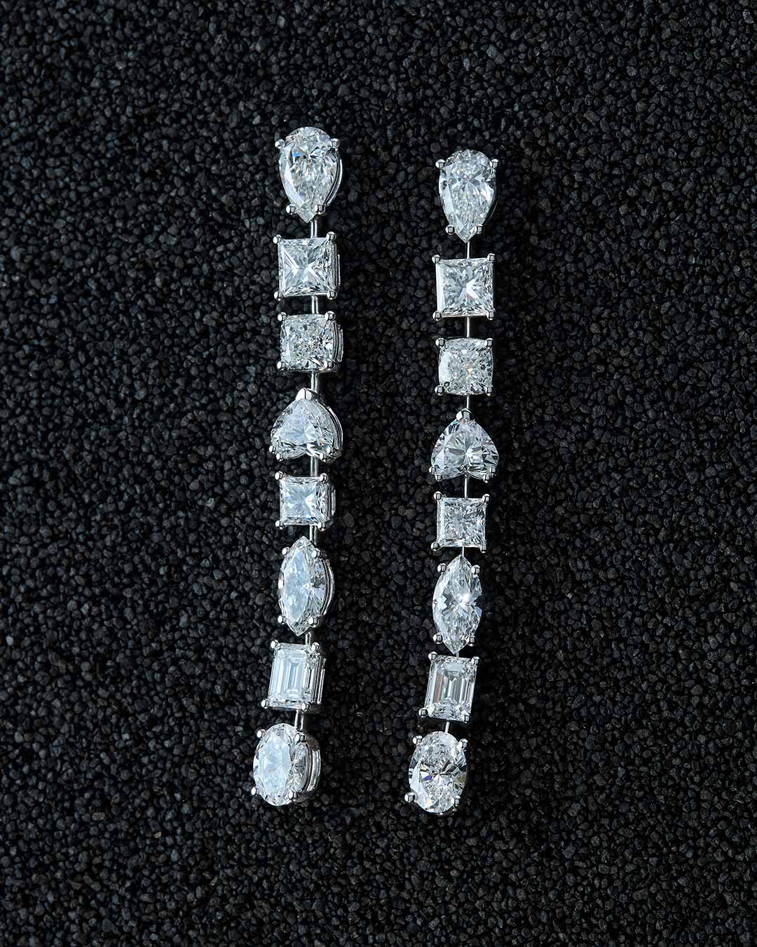 Pendientes largos sobre fondo negro texturizado. Cada pieza presenta una secuencia vertical de diamantes en distintos cortes. Engastados en oro blanco, los diamantes forman una composición simétrica que resalta la precisión del diseño y la sofisticación de la joya.